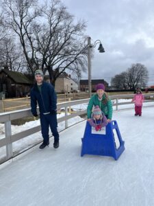 picture of a family skating. Man wearing blue coat and hat, a woman wearing a green coat and purple hat, and a child wearing a purple coat and hat using adaptive skate equipment.