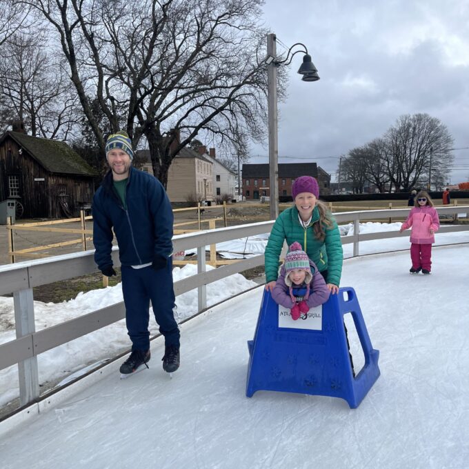 picture of a family skating. Man wearing blue coat and hat, a woman wearing a green coat and purple hat, and a child wearing a purple coat and hat using adaptive skate equipment.
