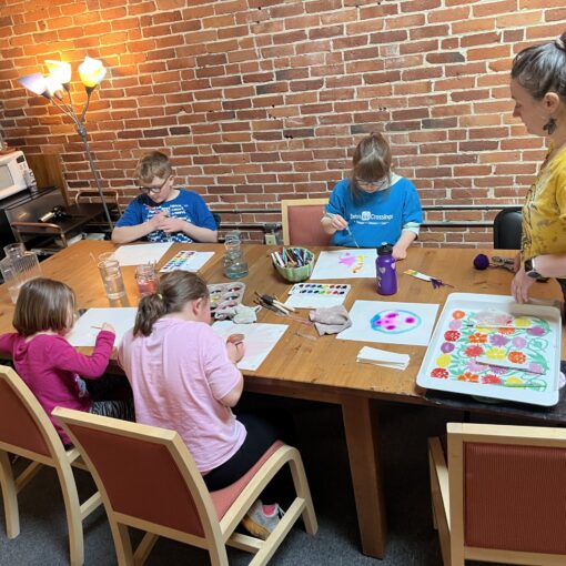 picture of 4 children and adult woman around a table painting on paper.